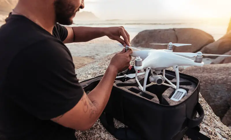 Young man sitting at rocky beach with a drone. Man setting up / dismantling a drone.