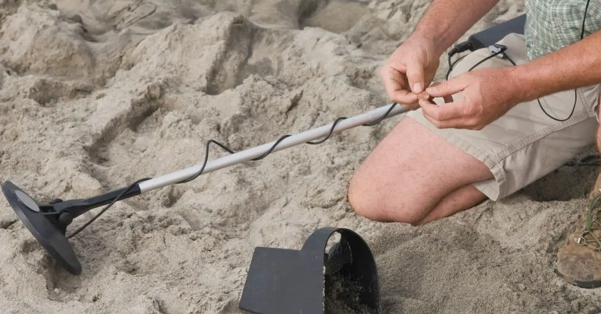 Man kneeling in the sand with a metal detector and a coin.